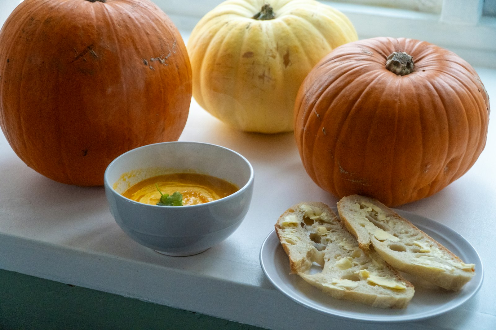 Pumpkin soup in a bowl next to whole pumpkins and bread on a windowsill