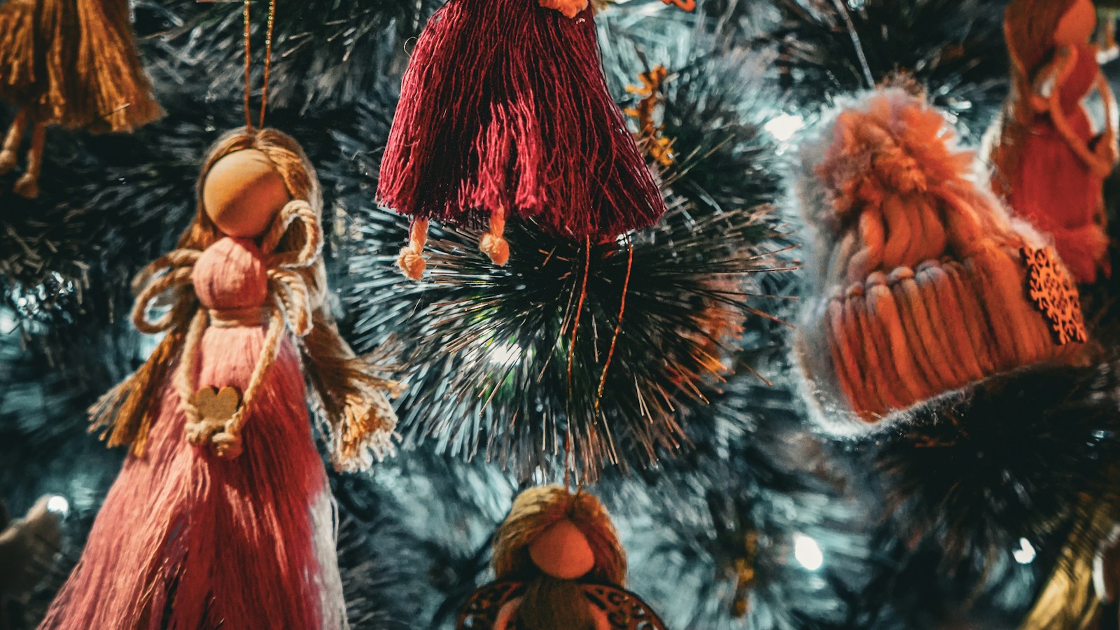 Handmade yarn angel ornaments in red and orange hanging on a dark green Christmas tree