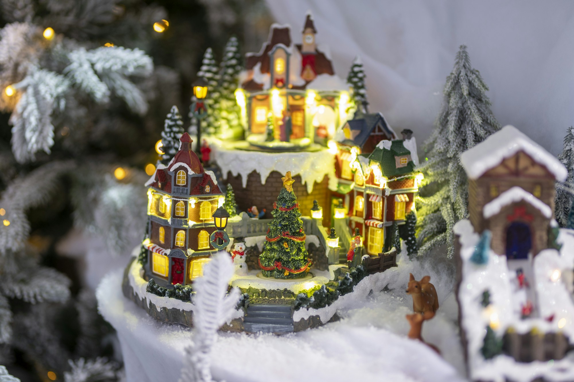 A miniature Christmas village glowing with warm lights next to snowy trees
