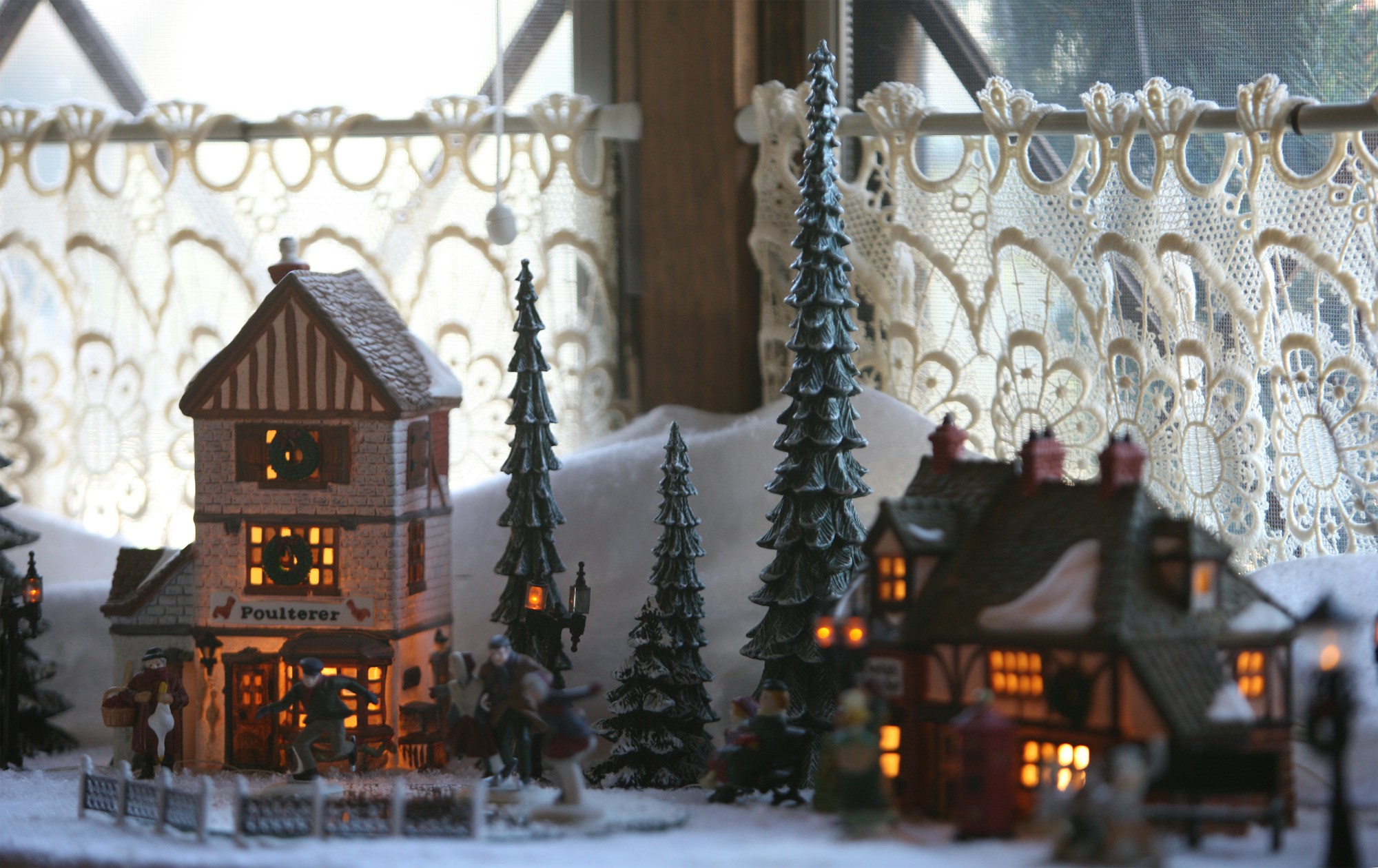 Miniature Christmas village houses and trees on a white snow blanket in front of a lace curtained window