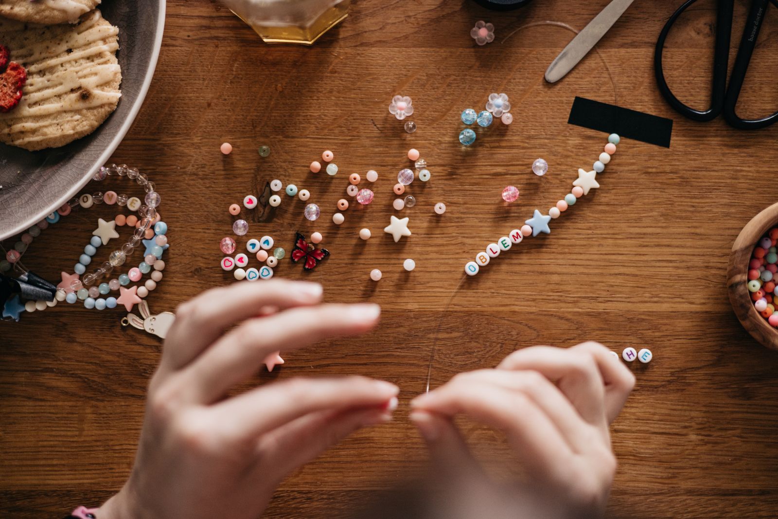 Hands threading colorful clay and letter beads onto a bracelet cord on a wooden table