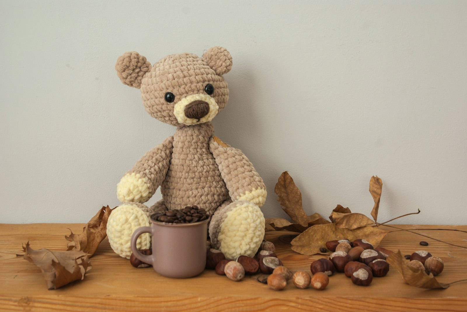 Crocheted amigurumi teddy bear sitting by a mug with autumn leaves