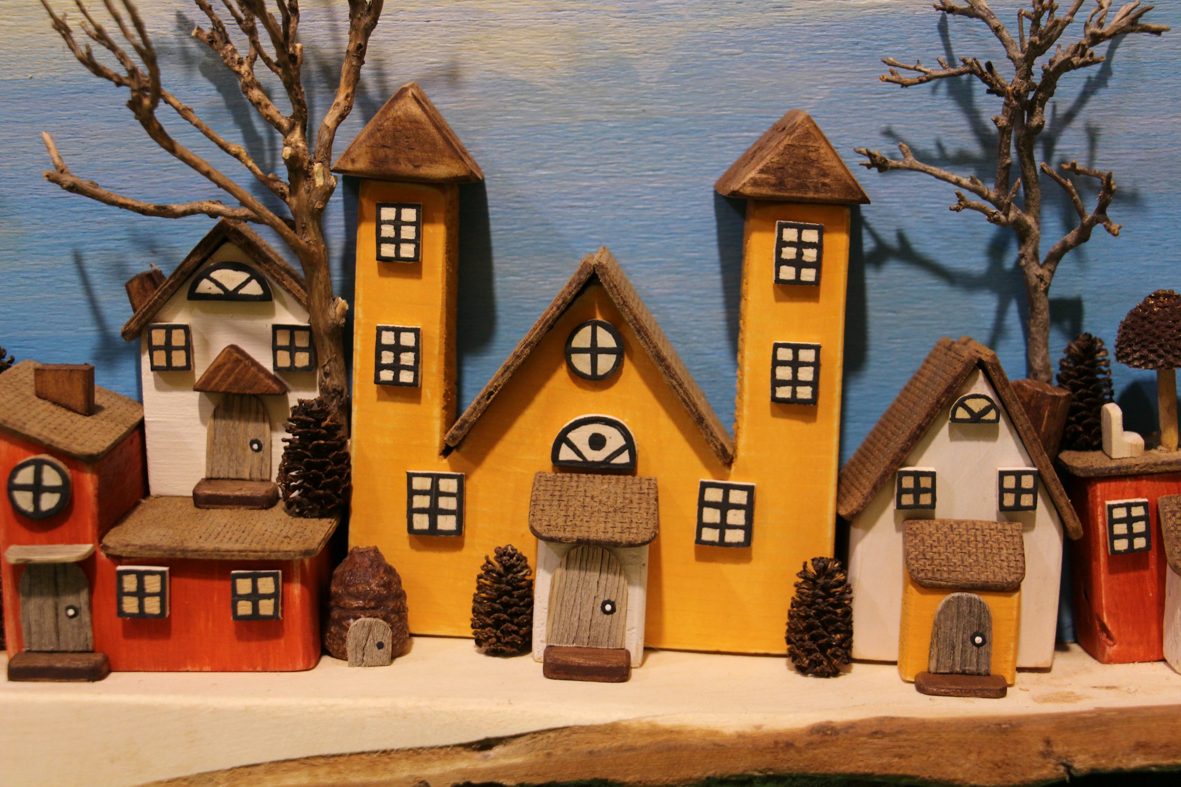 DIY Christmas village of painted wooden houses with pine cones and twig trees lined up on a shelf