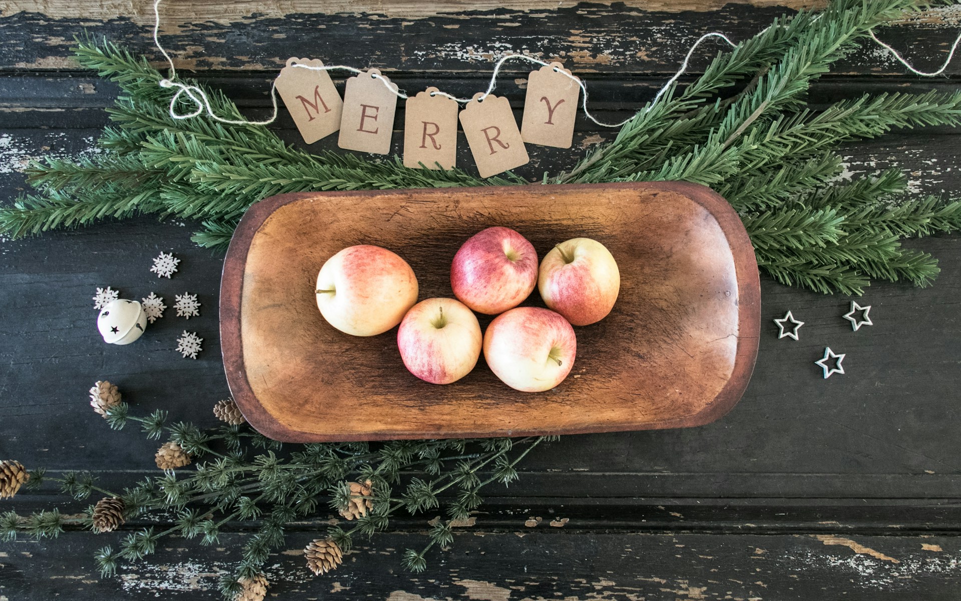 Five red apples in a wooden dough bowl surrounded by fresh evergreen on a dark wooden table