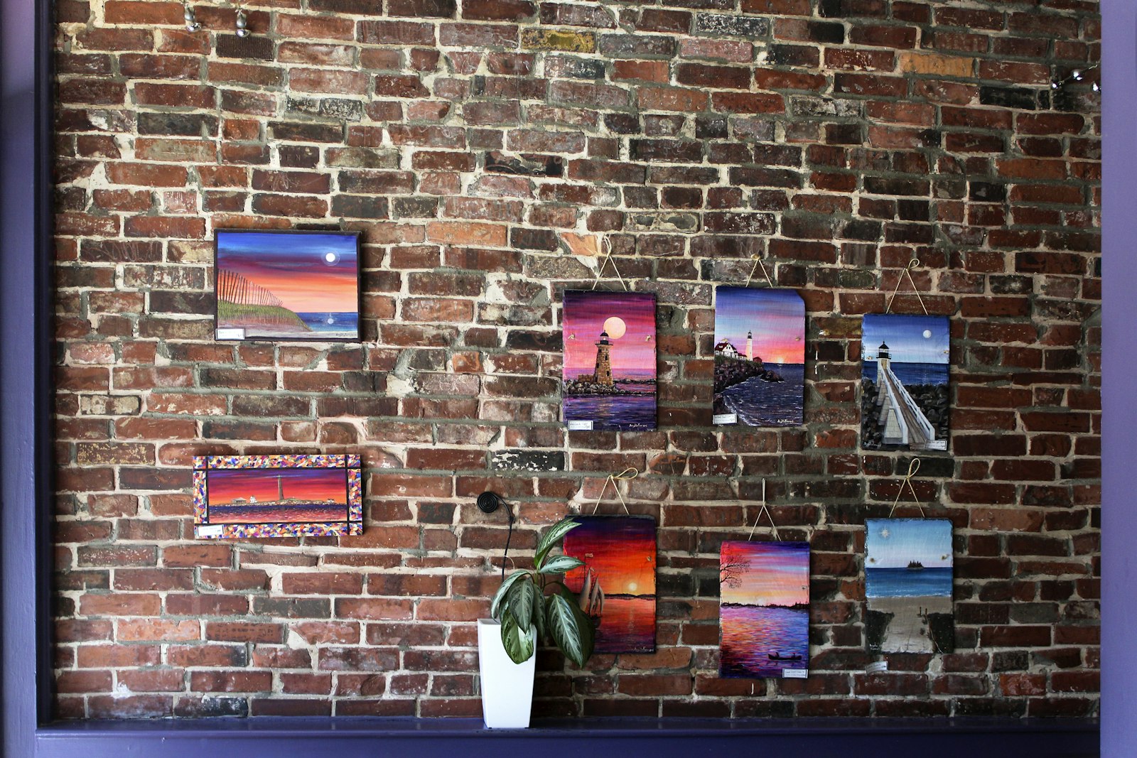 George W Bush painting inspiration: brick wall covered with small colorful canvas paintings of sunsets and lighthouses above a green plant