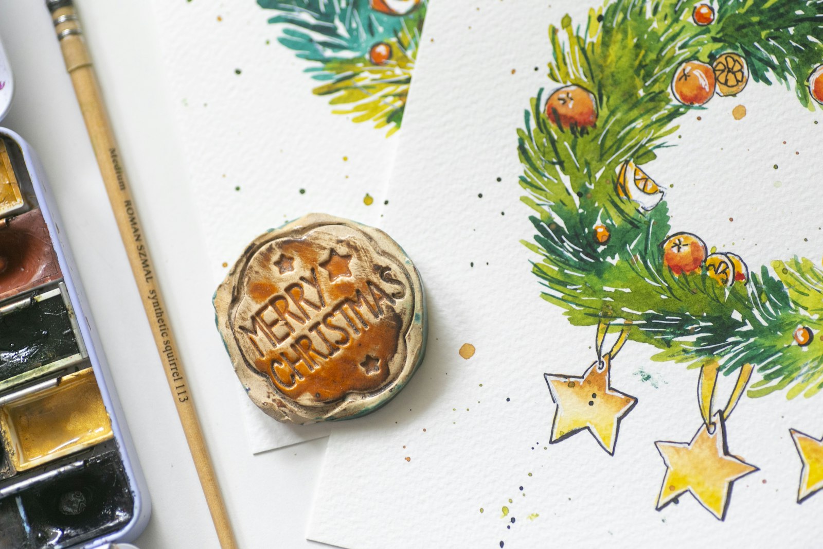 Hand painted watercolor Christmas wreath card beside a watercolor paint palette and brush