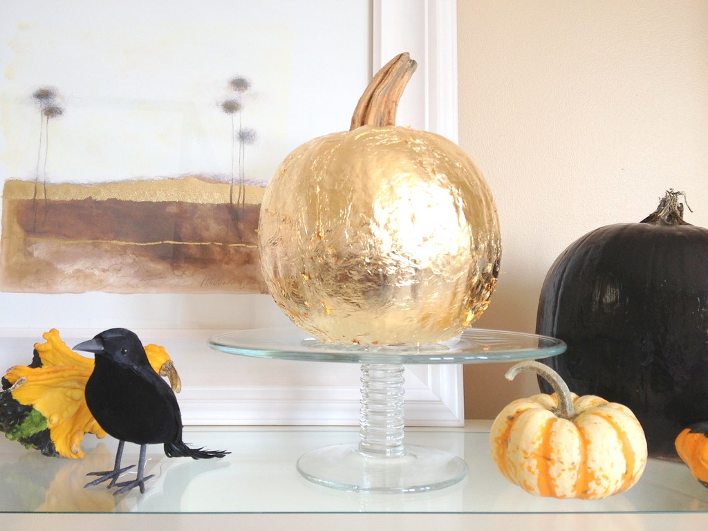 Shiny gold painted pumpkin on a glass cake stand next to a black pumpkin