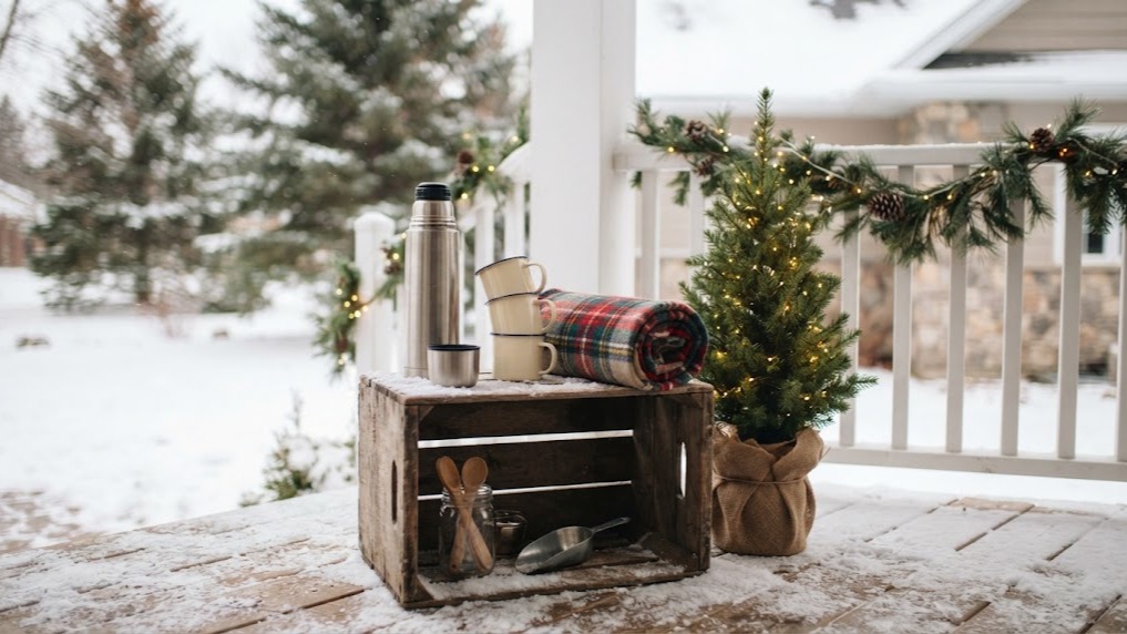 Hot cocoa bar ideas: a wooden crate porch cocoa station with a thermos, enamel mugs and a plaid blanket