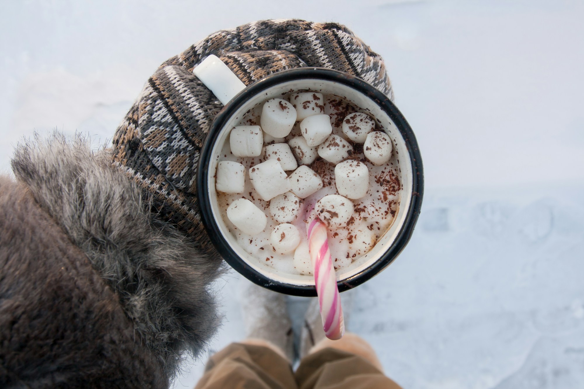 An enamel mug of hot cocoa with marshmallows and a candy cane held outdoors in the snow