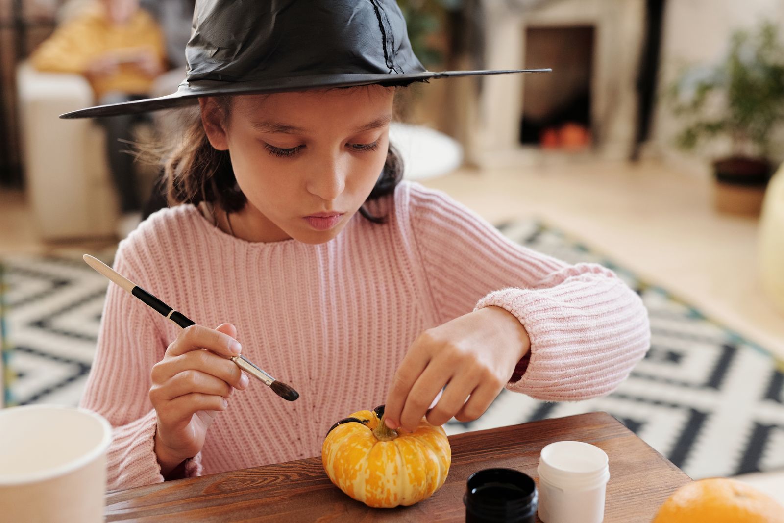 Young girl in a witch hat painting a small pumpkin at a craft table