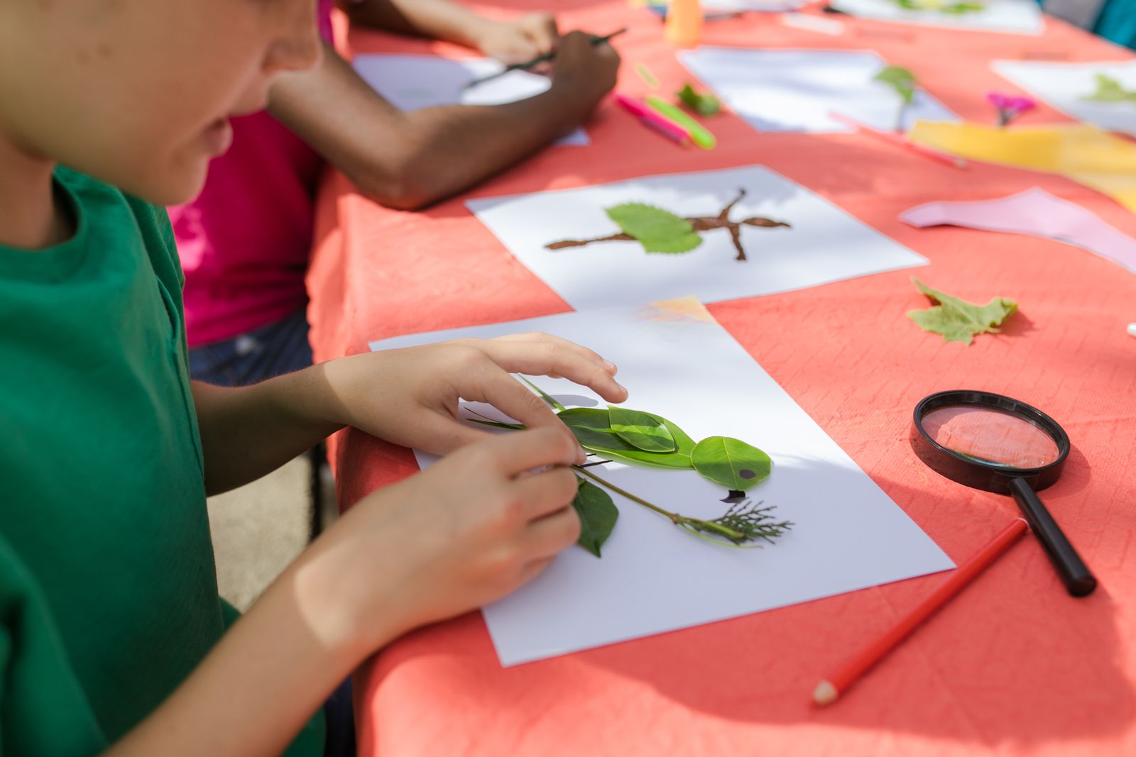 Child making a leaf creature collage on white paper at an outdoor craft table