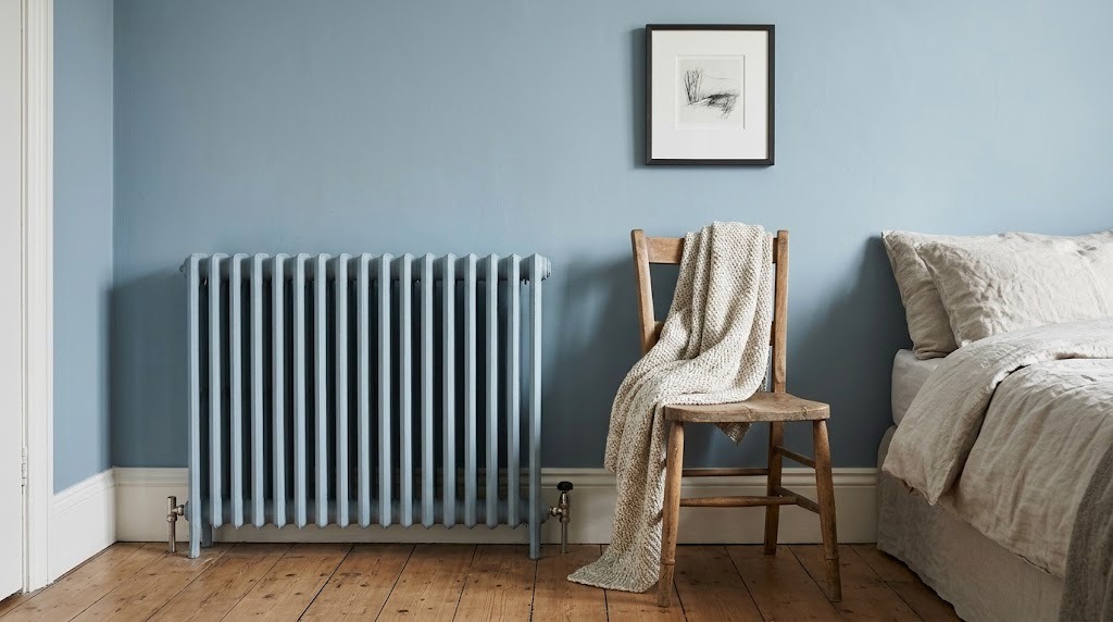 Old column heater painted the same soft blue as the bedroom wall beside a wooden chair