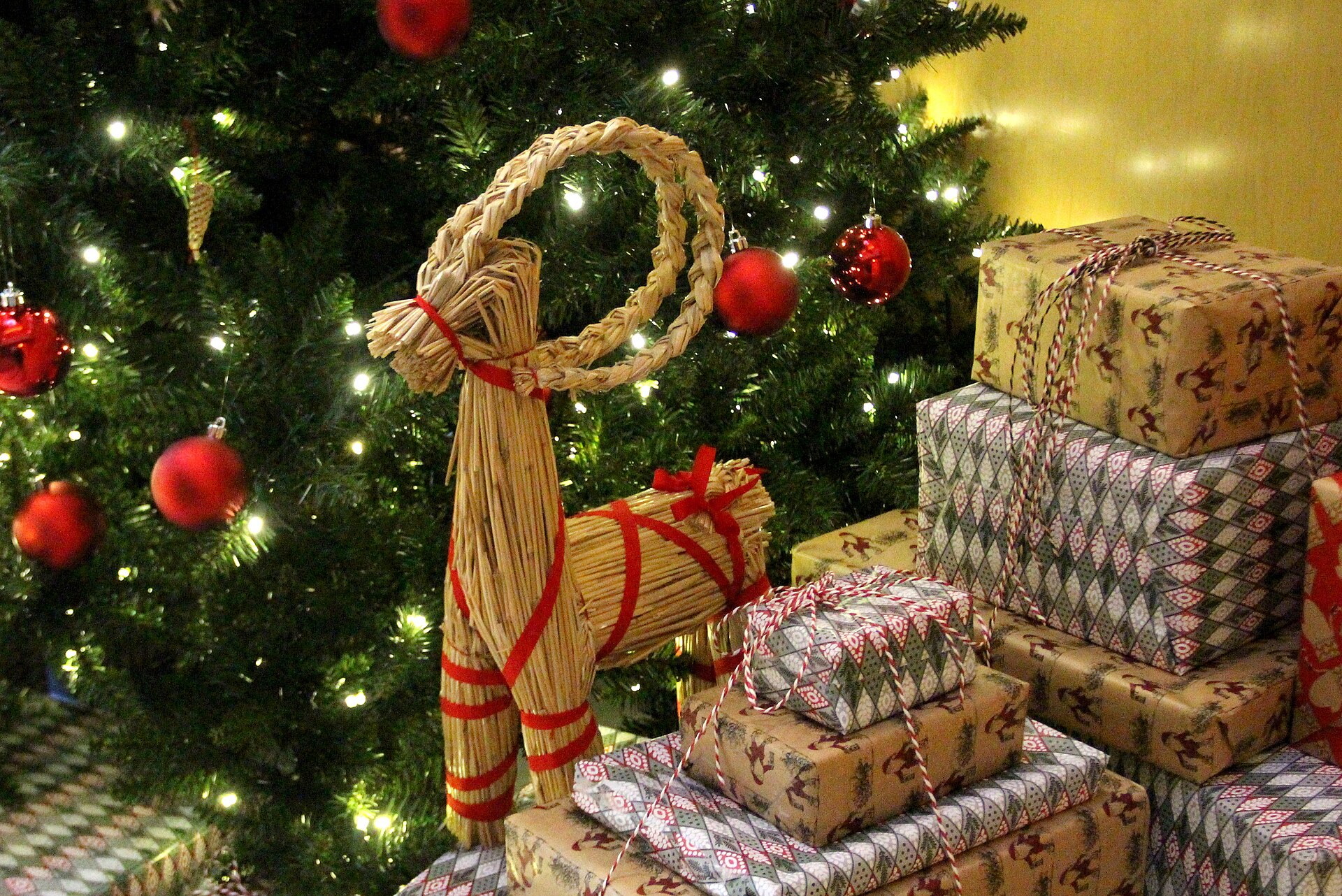 Swedish julbock straw goat tied with red ribbon beside a Christmas tree and gifts