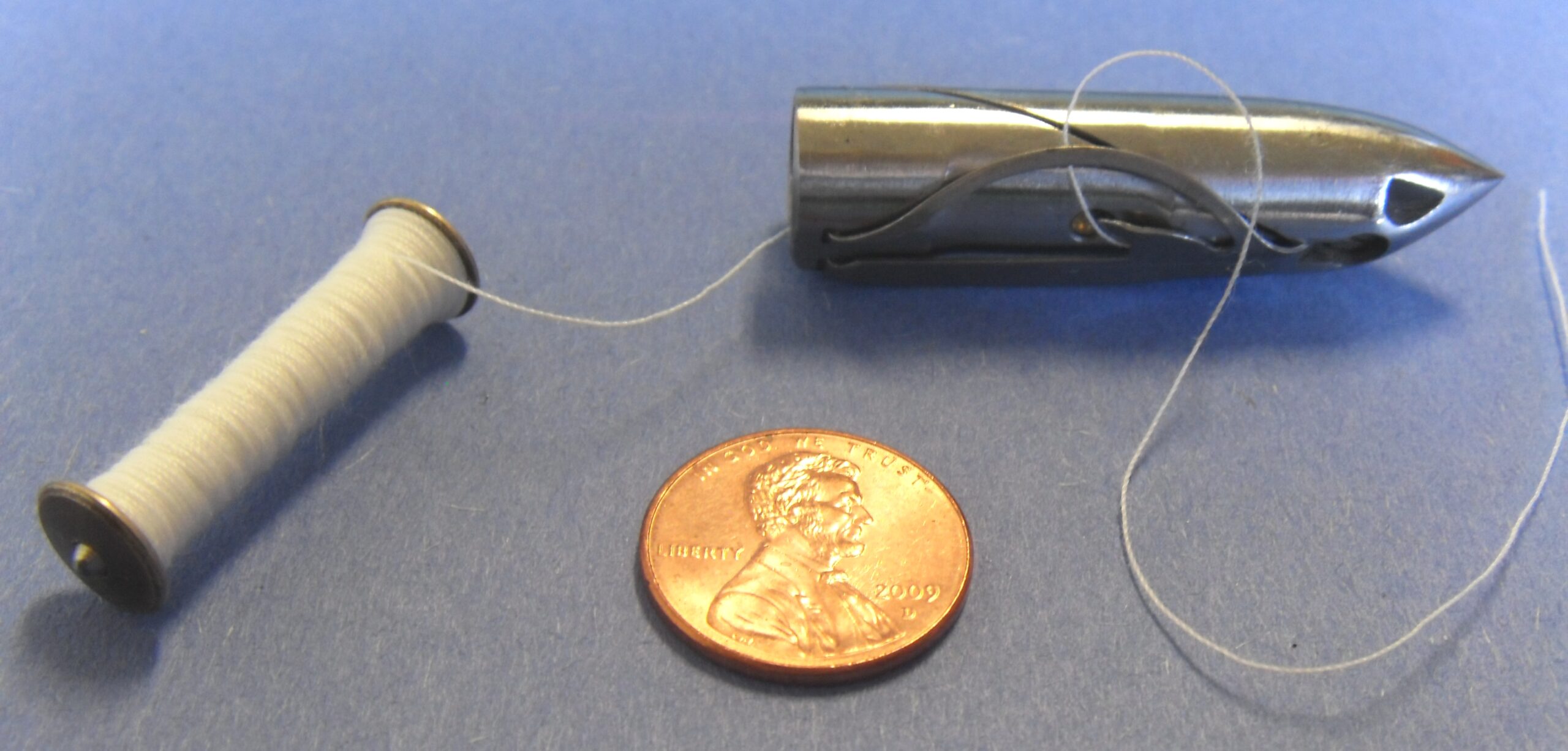 A long bullet shaped shuttle and bobbin from an antique sewing machine next to a penny