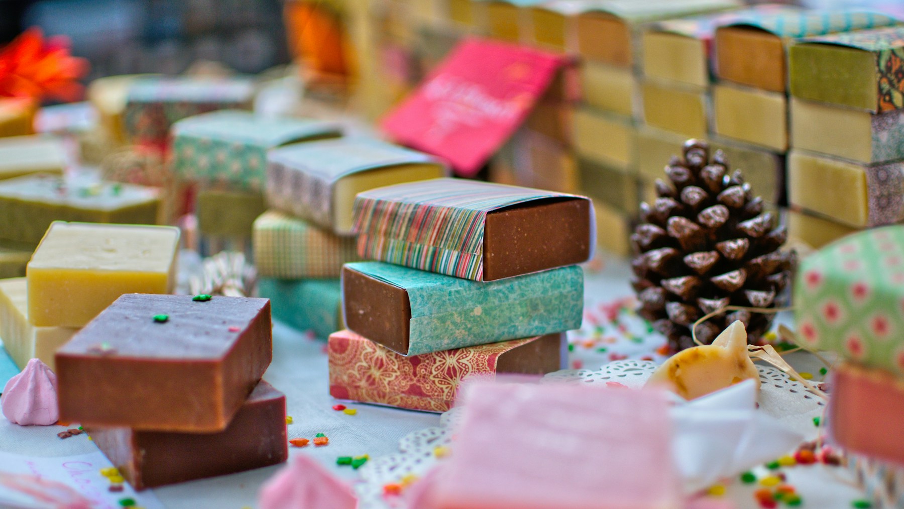 Handmade soap bars in patterned paper sleeves stacked on a market table with a pine cone