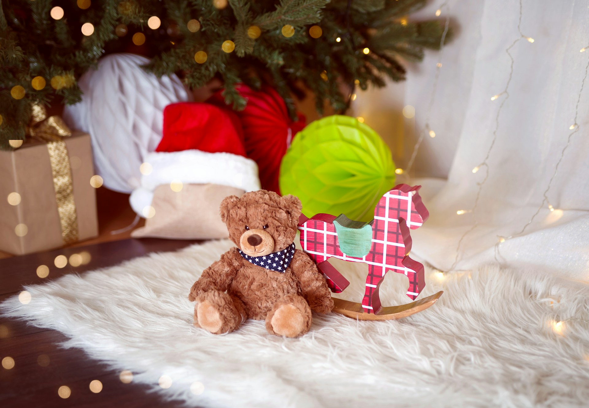 Brown teddy bear on a white fur throw next to a plaid rocking horse and gifts