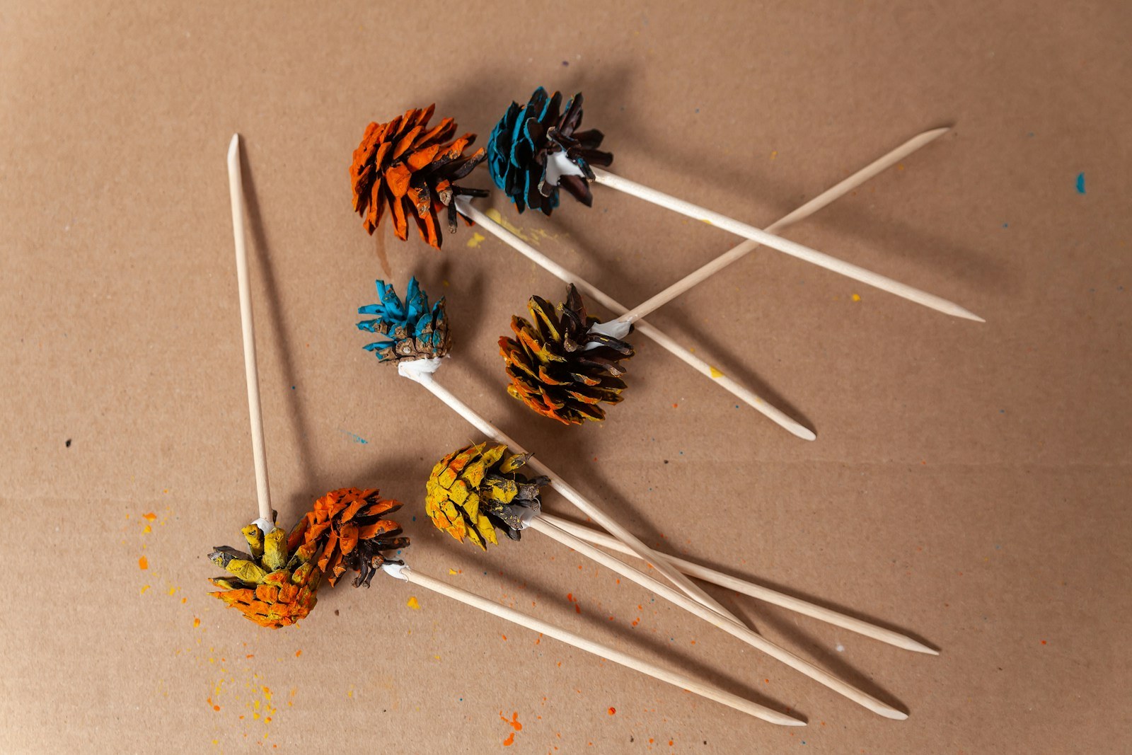Andy Warhol Christmas pine cones painted bright orange, turquoise and yellow drying on wooden sticks on brown paper