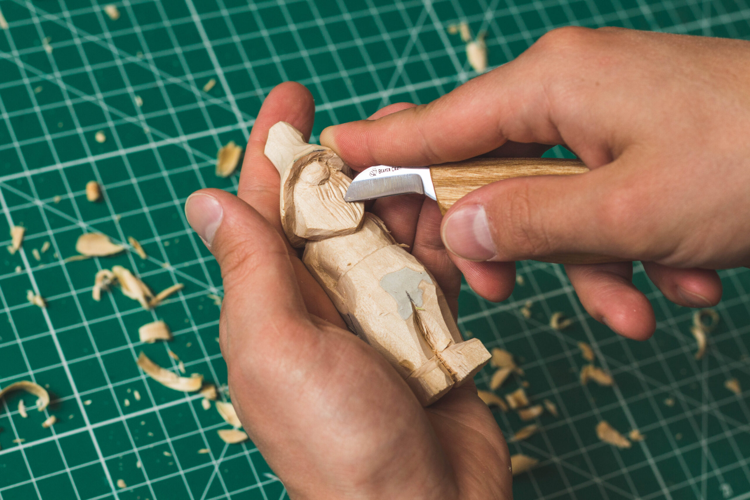 Hands whittling a small wooden figure with a knife over a cutting mat