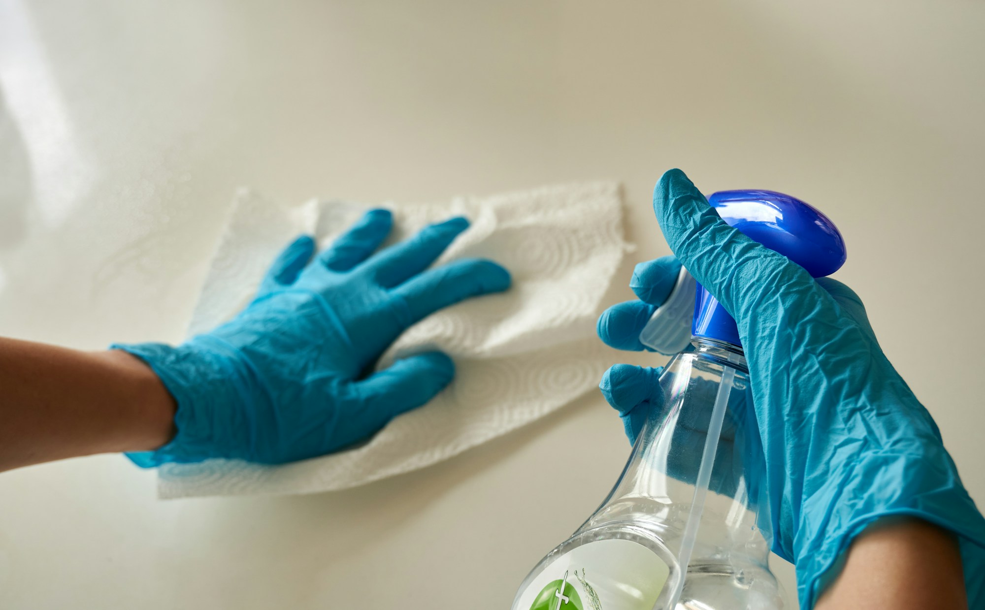 Gloved hands wiping a white counter with a soft cloth and a spray bottle