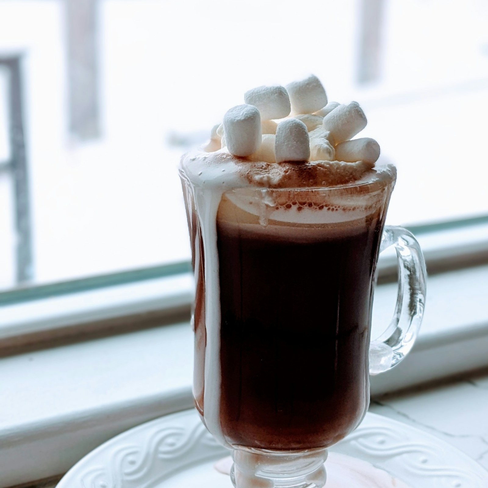 Glass mug of rich hot chocolate topped with marshmallows by a snowy window