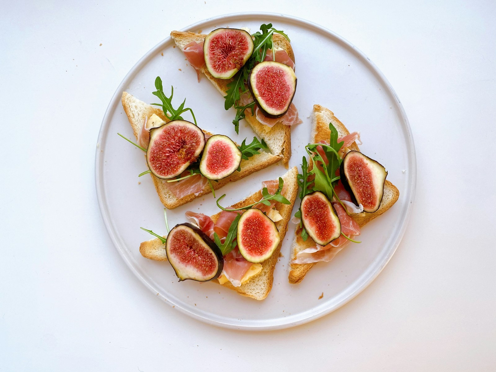 Toasts topped with fresh figs, prosciutto and arugula on a white plate
