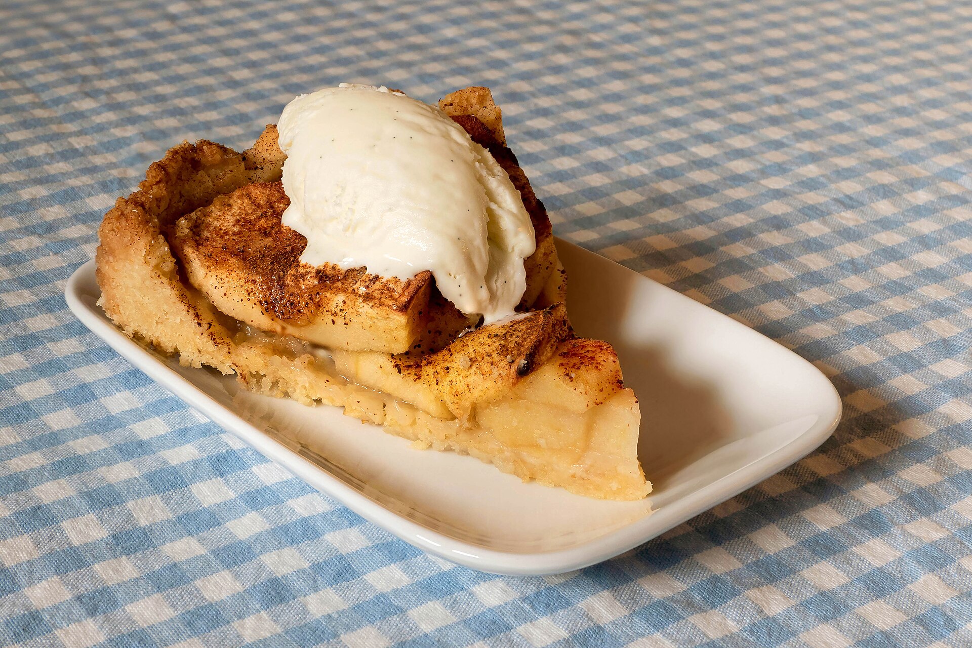 Slice of apple cake with vanilla ice cream on a blue gingham cloth