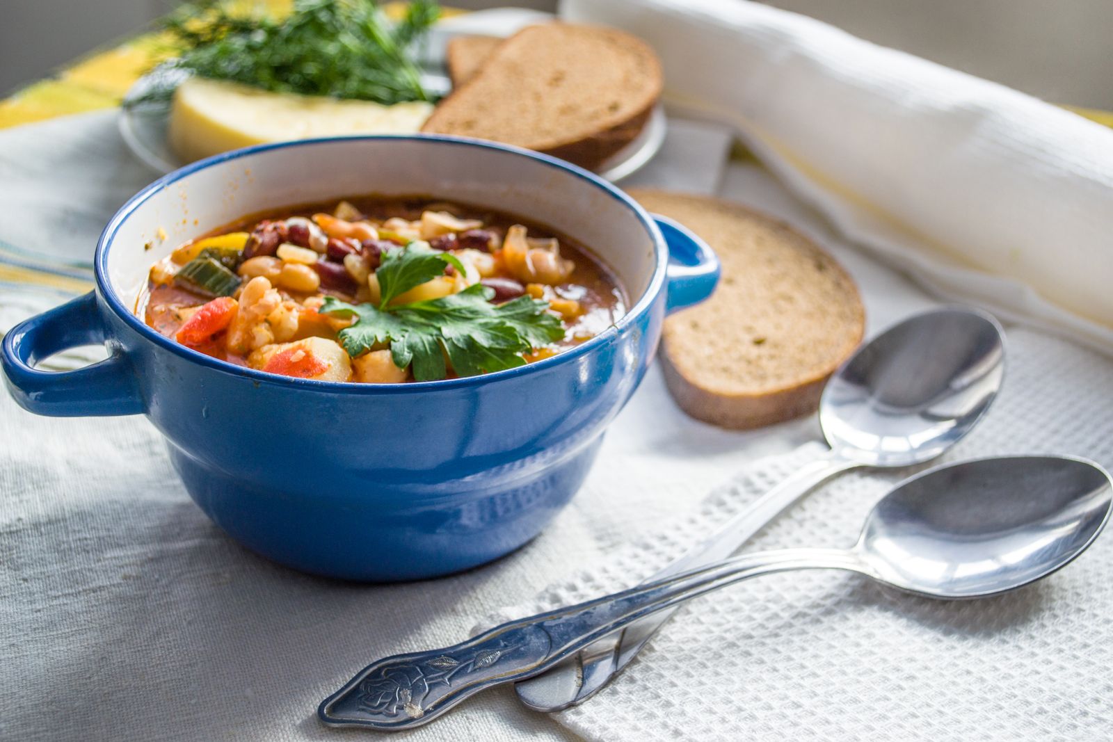 Vegetable and bean soup in a blue pot with bread and spoons