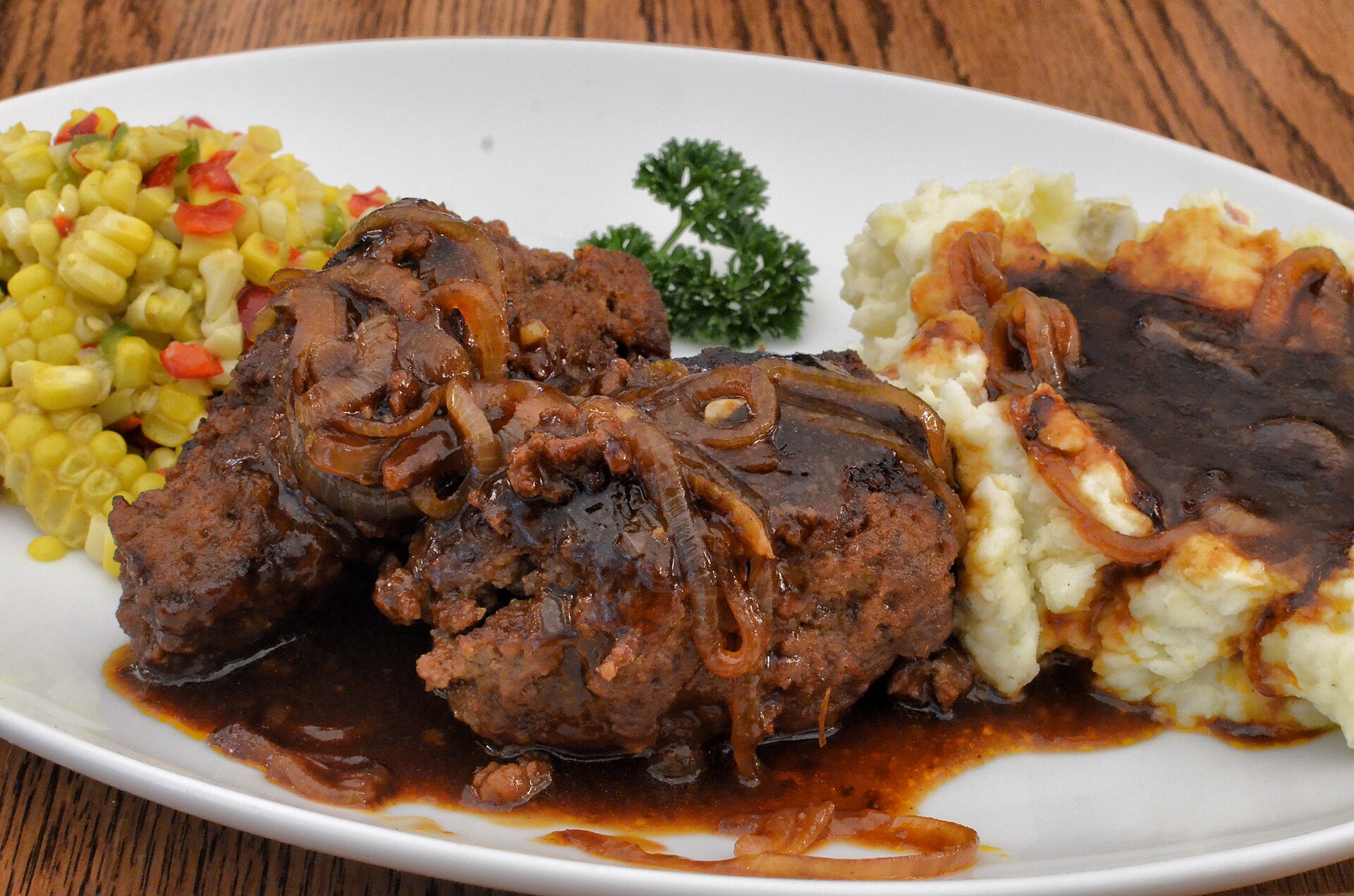 Salisbury steak with brown onion gravy, mashed potatoes and corn on a white plate