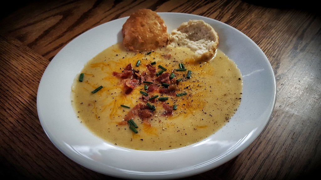 White bowl of creamy cheesy potato soup topped with bacon and green onion, with a biscuit