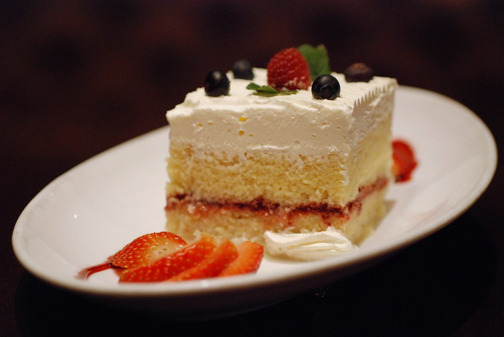 Square of tres leches with whipped cream and strawberries on a white plate