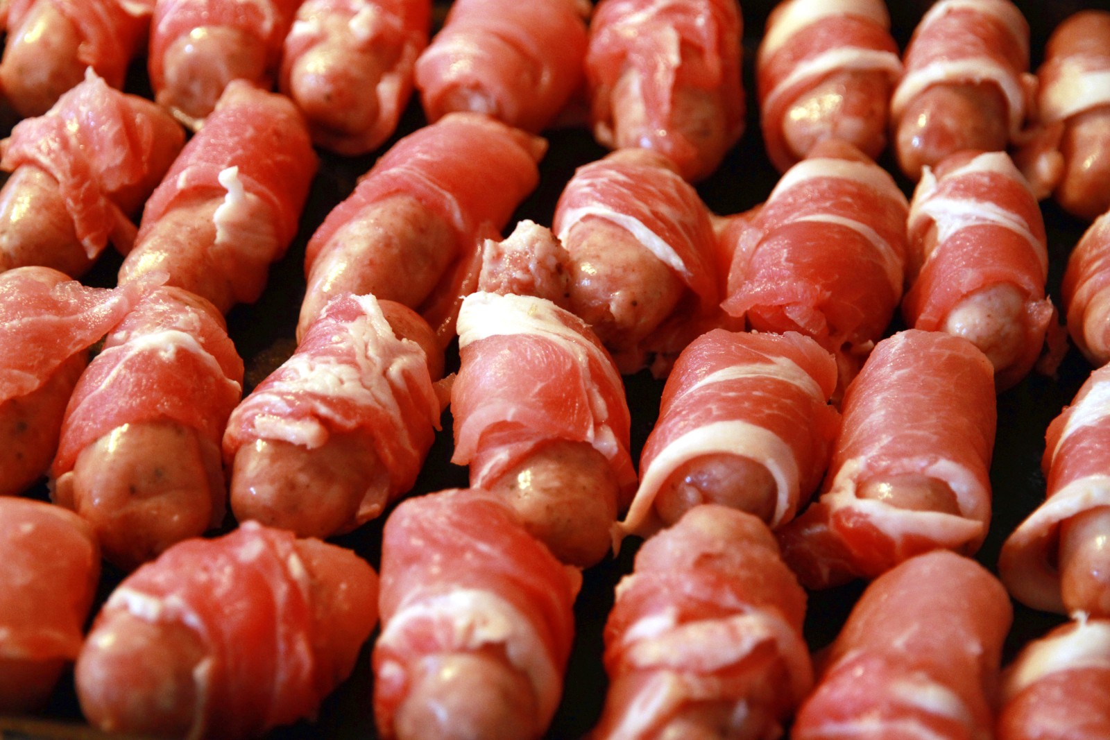 Small sausages wrapped in crispy bacon on a baking tray