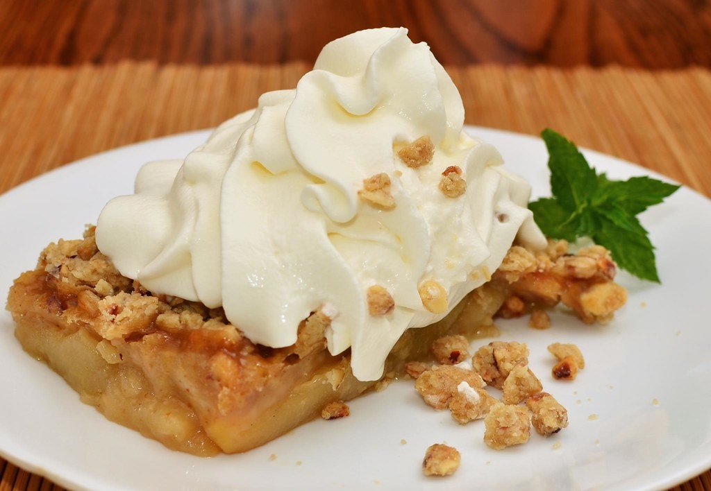 Apple crisp topped with whipped cream and chopped nuts on a white plate